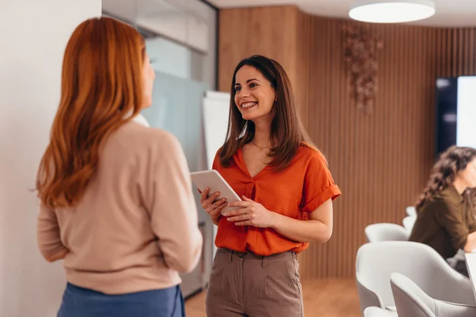 Two young adult businesswomen engaging in a lively discussion in a modern office, with one holding a digital tablet and smiling, showcasing teamwork and collaboration