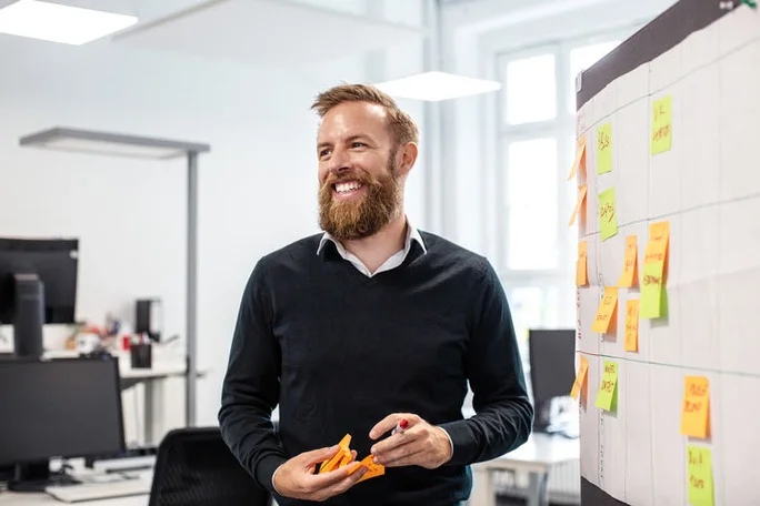 Confident bearded businessman looking away while standing by bulletin board in creative office